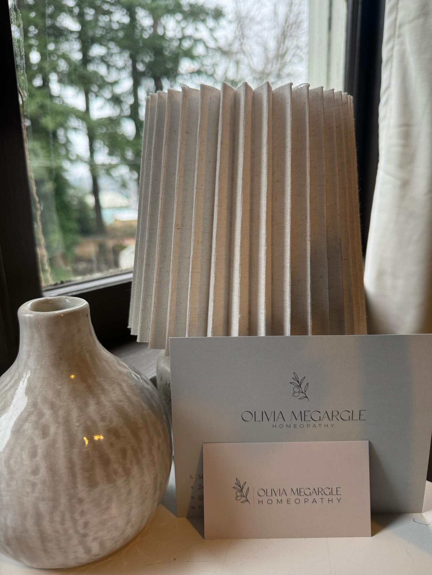 Olivia Megargle Homeopathy cards on a windowsill, evergreens beyond the glass